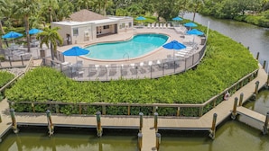A pool area with a circular pool, lounge chairs, umbrellas, and a wooden deck over water.