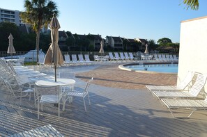 Un área junto a la piscina con sillones de descanso y mesas, una piscina y un edificio al fondo.