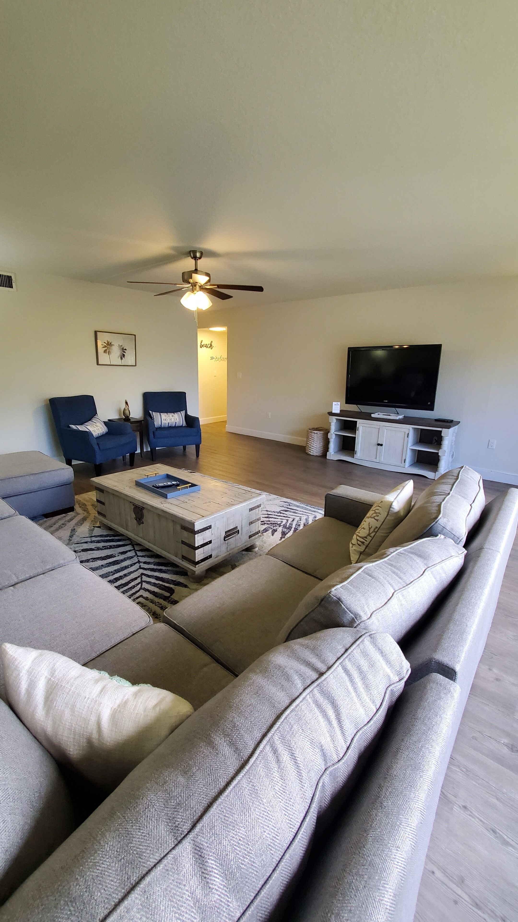A living room with a large sectional sofa, a coffee table, two armchairs, a TV, and a ceiling fan.