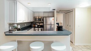Una cocina moderna con armarios blancos, electrodomésticos de acero inoxidable y una encimera oscura. Hay dos taburetes blancos en la isla.