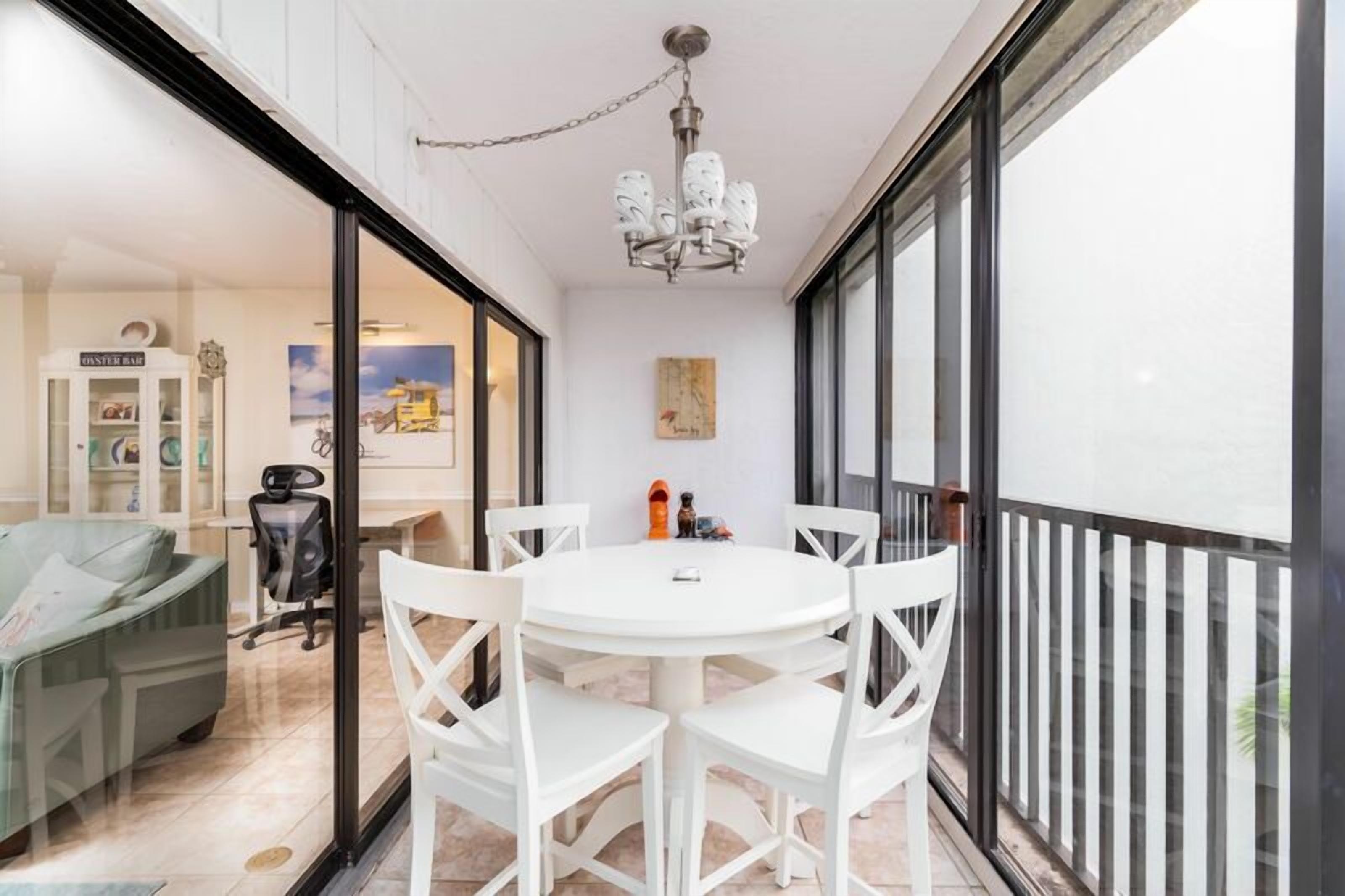 A modern dining area with a round table and white chairs, a chandelier, and a view through large glass doors to an adjacent room with a sofa and a cabinet.