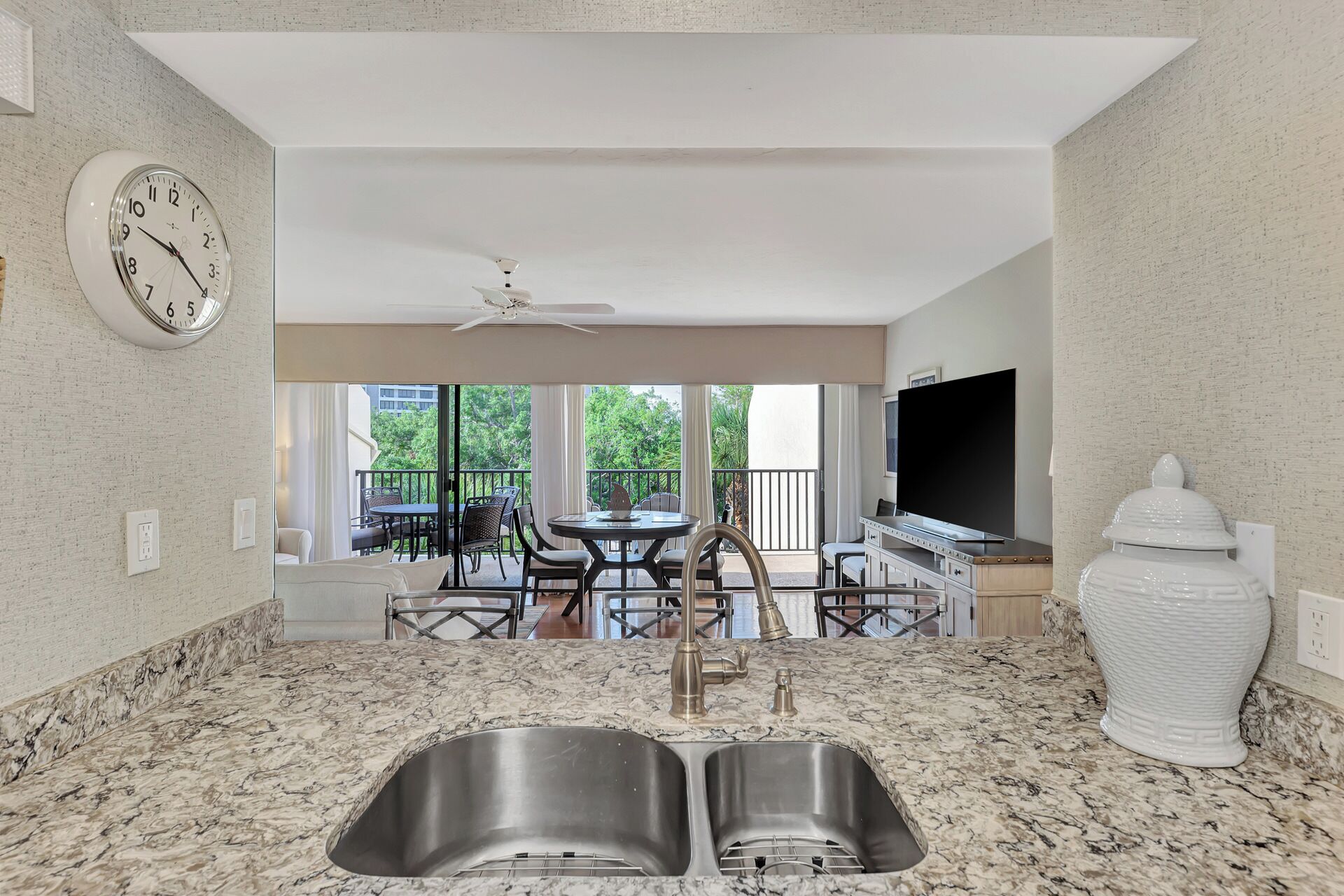A kitchen with a double sink, a dining area with a table and chairs, a ceiling fan, and a large window overlooking a balcony with outdoor furniture.
