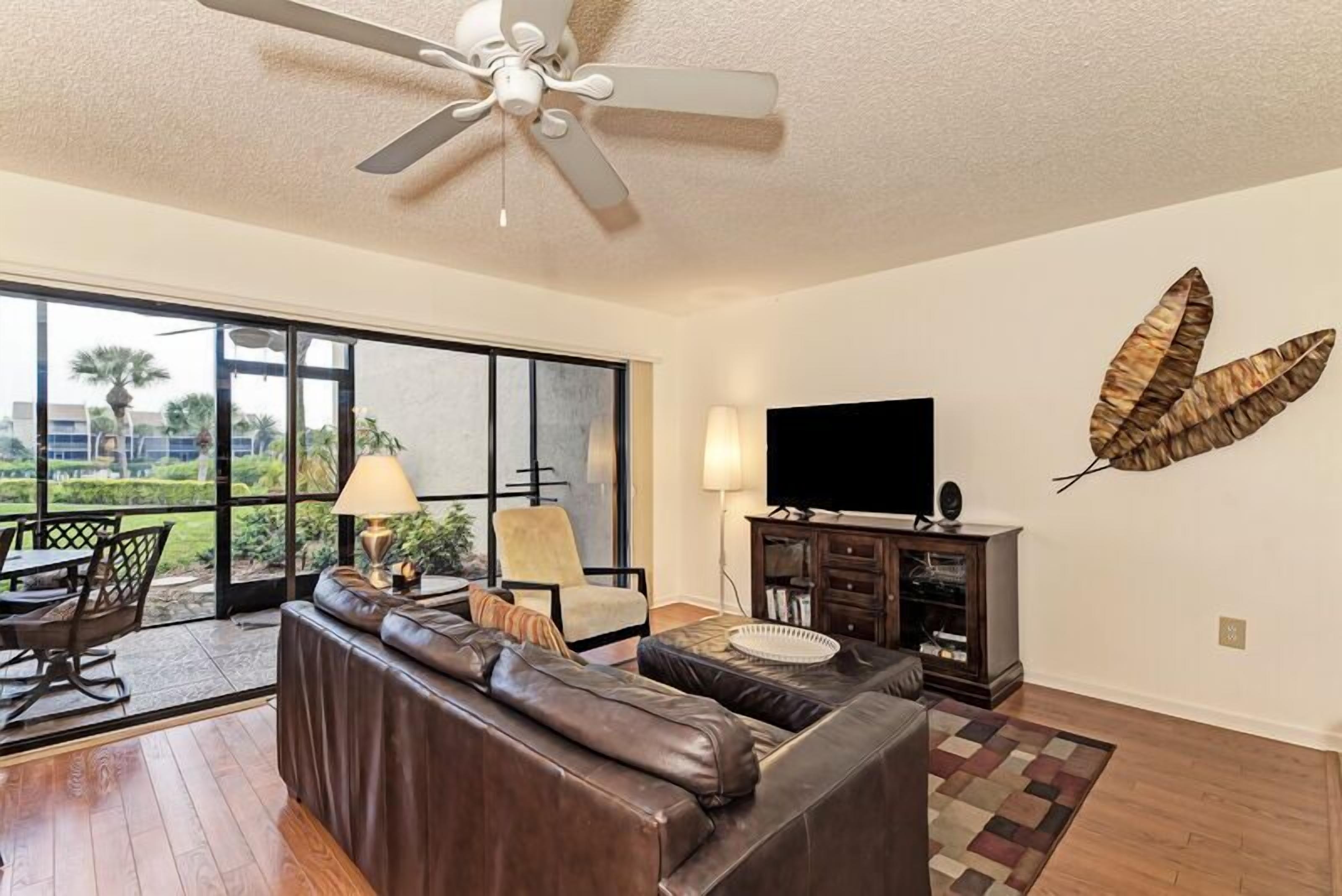 A living room with a leather sofa, a TV, a ceiling fan, and a large window with a view of palm trees.