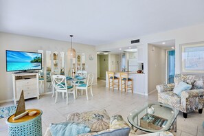 A spacious living area with a television, dining table, and chairs, a kitchen with a bar, and a patterned armchair. - Midnight Cove II 633F - Serene Vacation Rental on Siesta Key (Siesta Key)