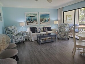 A living room with a white sofa, two patterned armchairs, a glass coffee table, and artwork on the walls.