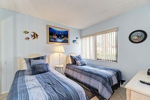 A bedroom with two beds, a nightstand, a desk, and a window with blinds. - Midnight Cove II 810F - Roomy Bayside Condo in Siesta Key (Siesta Key)