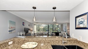 A modern kitchen with granite countertops, a sink, and pendant lighting. There is a dining area with a table and chairs visible through large windows. - Midnight Cove II 820F - Updated Siesta Key Bayside Rental, Pool and Tennis Acces (Siesta Key)