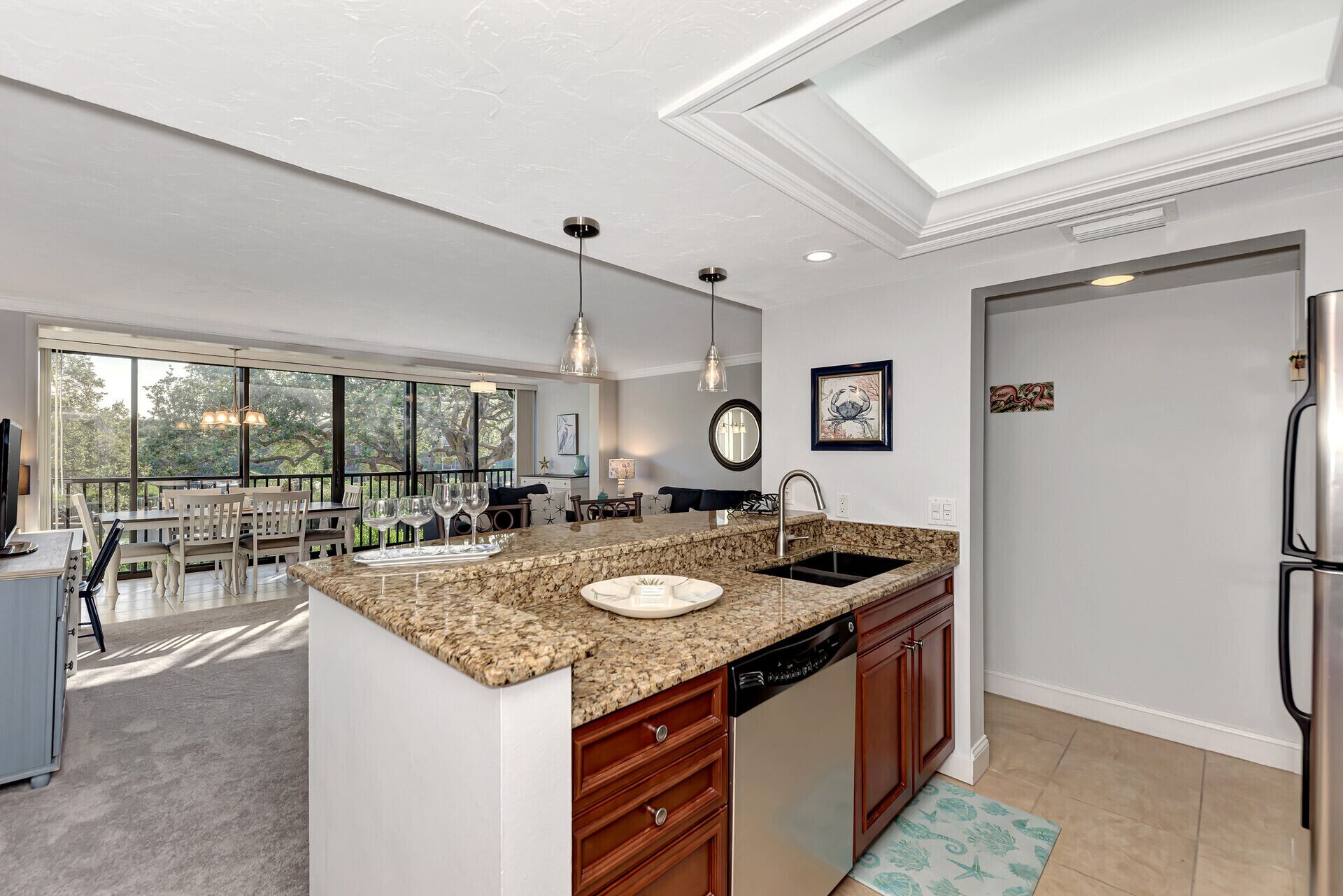 A modern kitchen with granite countertops, a dishwasher, and a large island. There is a dining area with a view of trees through large windows.