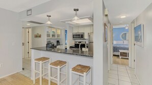 A modern kitchen with white cabinets, a central island with bar stools, a microwave, and a refrigerator. - Midnight Cove II 822F - Relaxing Vacation Rental in Siesta Key (Siesta Key)