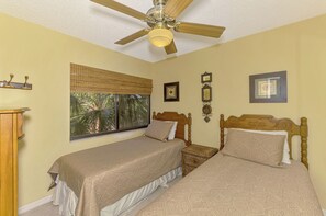 Une chambre avec deux lits simples, un ventilateur de plafond, une fenêtre donnant sur des palmiers et des cadres photo accrochés au mur.