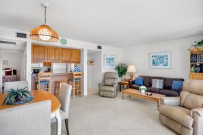 A modern living room with a sofa, coffee table, and a dining area with chairs and a table. There is a kitchen area with wooden cabinets and a hanging light fixture. - Midnight Cove II 833F - Siesta Key Condo with Balcony and Marina View! (Siesta Key)
