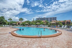 Una piscina con sillas de descanso y sombrillas, rodeada de palmeras y un edificio de hotel al fondo.