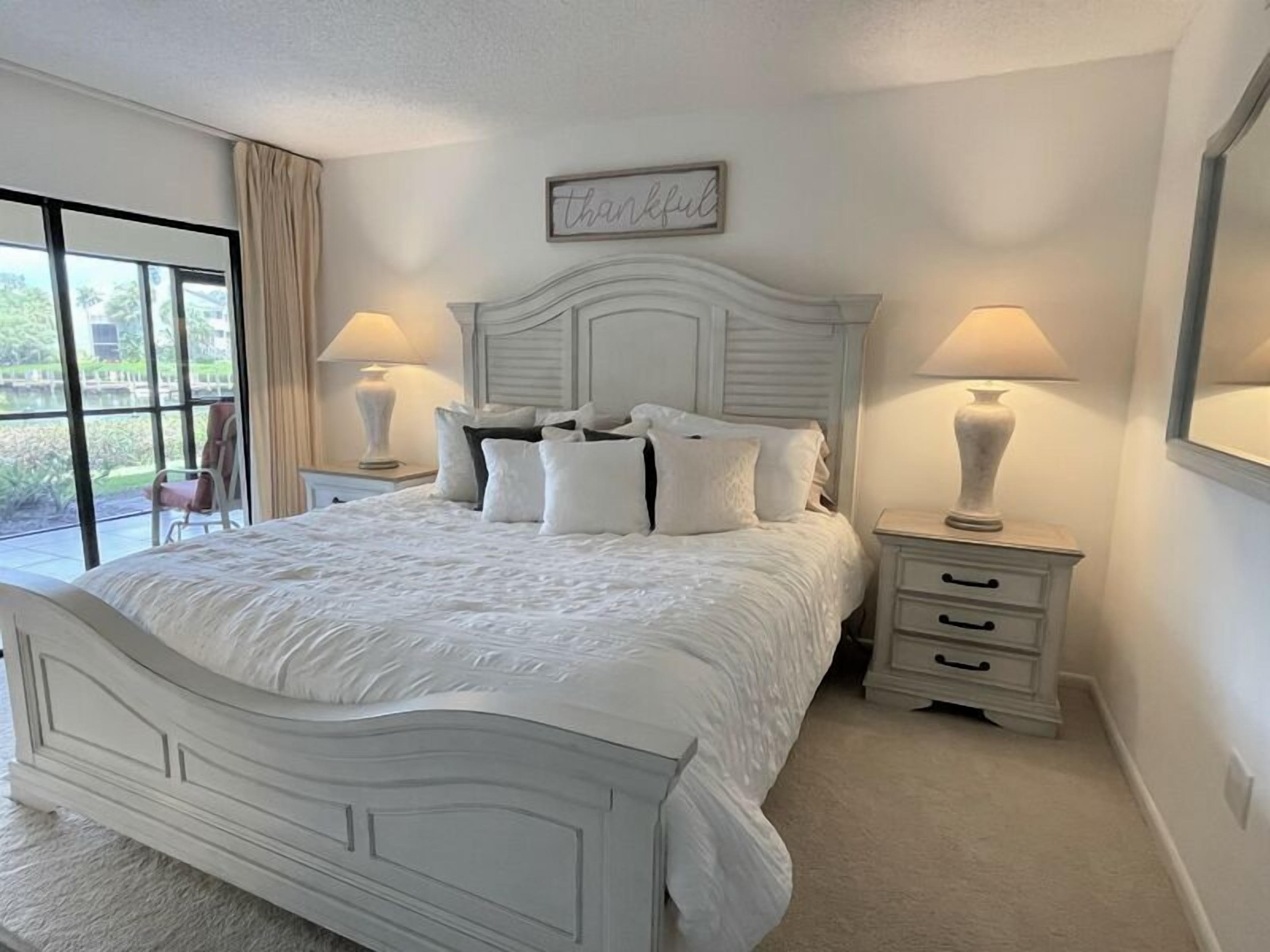 A bedroom with a large bed, two bedside tables, a window with curtains, and a wall-mounted sign.
