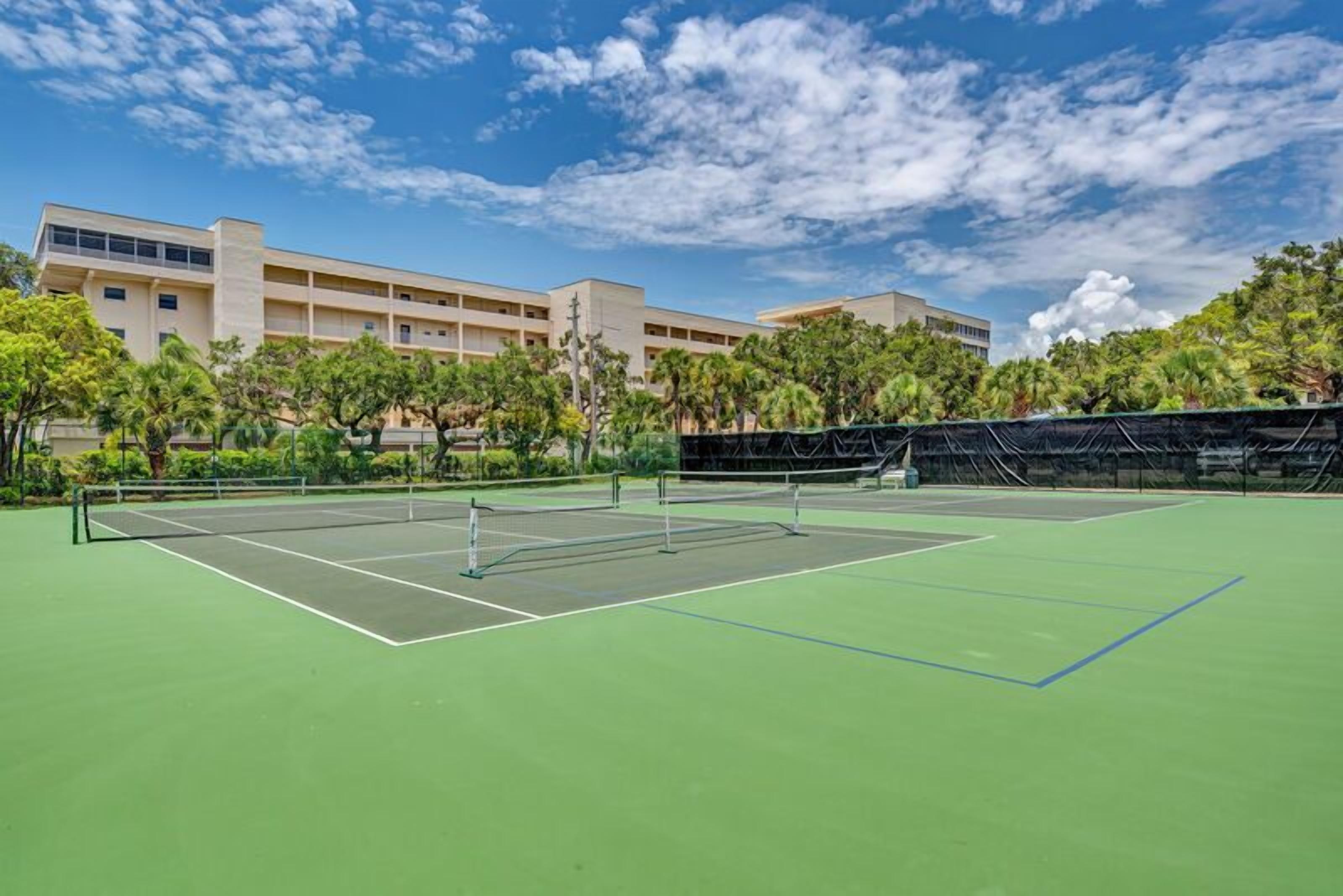 A tennis court with a net in the center, surrounded by trees and a building in the background.