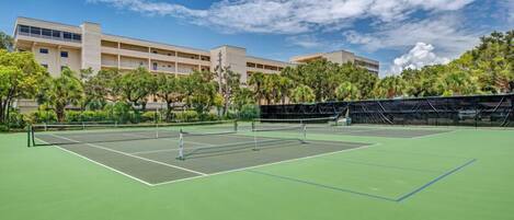 A tennis court with a net in the center, surrounded by trees and a building in the background.