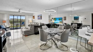 Ampla sala de estar com mesa de jantar de vidro, cadeiras cinzas, sofá e varanda com vista.