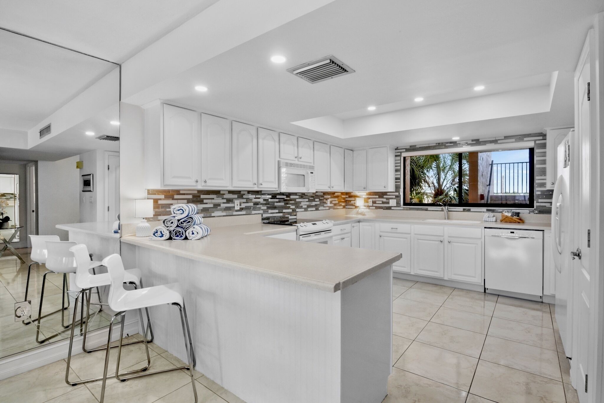 A modern kitchen with white cabinetry, a central island with bar stools, and a view of the outdoors.