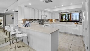Cozinha moderna com armários brancos, uma ilha central com banquetas e vista para o exterior.