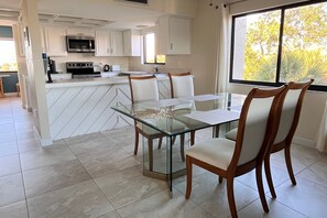 A modern kitchen with white cabinetry, a glass island with chairs, and a large window with a view of trees.
