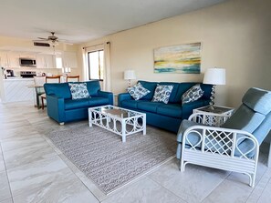 A modern living room with a blue sofa set, white coffee table, and a white armchair. A kitchen is visible in the background.