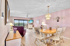A dining area with a wooden table and white chairs, a ceiling fan, and a TV in the background.