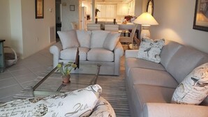 A living room with a sofa set, a coffee table with a plant, and a lamp. There is a kitchen area in the background. - Midnight Cove II 944C - Exceptional Water Views on Siesta Key (Siesta Key)