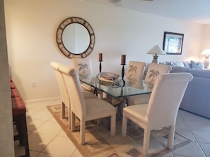 Un coin repas avec une table en verre entourée de quatre chaises, un miroir rond accroché au mur et un tableau encadré au-dessus d’un canapé.