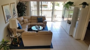 Un salon avec un canapé d’angle beige, une table basse ronde et un lampadaire. Une porte-fenêtre coulissante donne accès à un espace extérieur comprenant une terrasse et une vue sur l’extérieur.