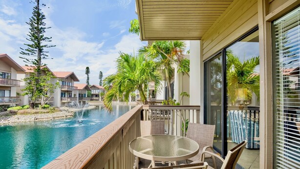 Un balcon doté d’une table et de chaises en verre, offrant une vue sur une piscine et un espace paysager agrémenté de palmiers et d’une petite cascade.