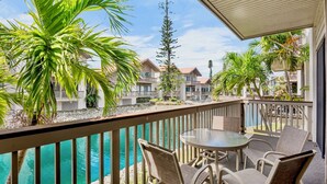 Un balcon avec une table et des chaises donnant sur une piscine, entouré de palmiers et d’un ensemble d’immeubles.