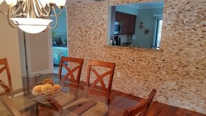 Un comedor con mesa de vidrio, sillas de madera y un bol de aperitivos. Una pared de ladrillo con un espejo refleja una cocina y un salón.