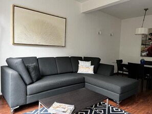 A modern living room with a sectional sofa, a coffee table, and a dining area in the background.