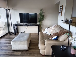 A living room with a flat-screen TV, a sofa, an ottoman, a coffee table, a floor lamp, a potted plant, and a wall-mounted shelf with decorative items.