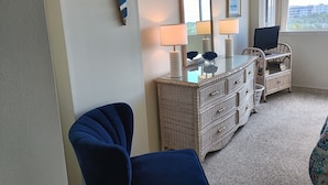 Ein Zimmer mit einem großen Spiegel, zwei Wandlampen, einer Korbmöbel-Kommode, einem Fernseher auf einem Tisch und Blick durch ein Fenster nach draußen.
