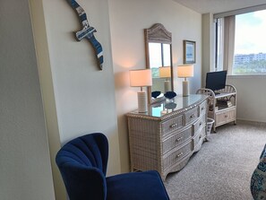 Una habitación con un espejo grande, dos lámparas de pared, un aparador de mimbre, un televisor sobre un mueble y vistas al exterior a través de una ventana.