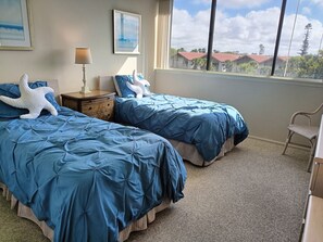 A bedroom with two beds, a nightstand, a lamp, and a window with a view of buildings.