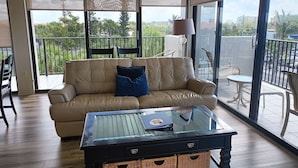 Ein Wohnzimmer mit einer Couch, einem Glastisch, Korbwannen, einem Balkon mit Blick auf Grünflächen und Gebäude.