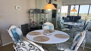 A dining area with a round table, chairs with patterned cushions, a decorative metal stand with glassware, and a view of the outdoors through large windows. - Our House at the Beach E-402 Beach view (Siesta Key)