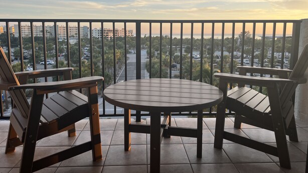A balcony with two wooden chairs and a round table, overlooking a cityscape with buildings and a road. - Our House at the Beach E-404 Sugar Sands Beach View!! (Siesta Key)