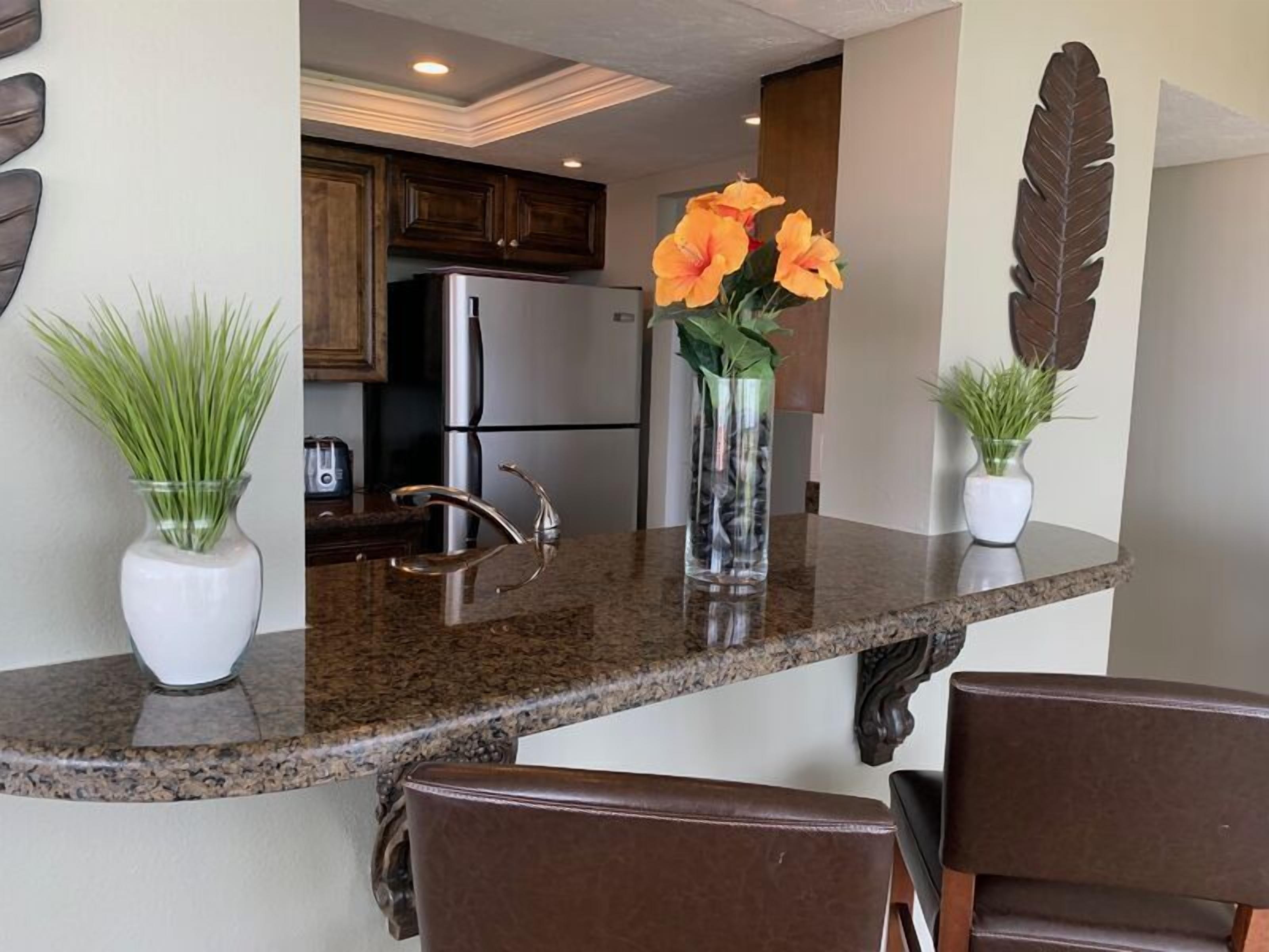 A kitchen with granite countertops, a stainless steel refrigerator, and wooden cabinets. There are two potted plants on the counter.