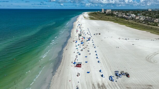 A wide beach with clear blue waters, a long stretch of white sand, and a few buildings in the background.