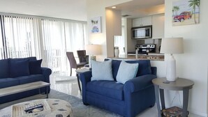 A living room with a blue sofa, a coffee table, and a dining area with a kitchen in the background.