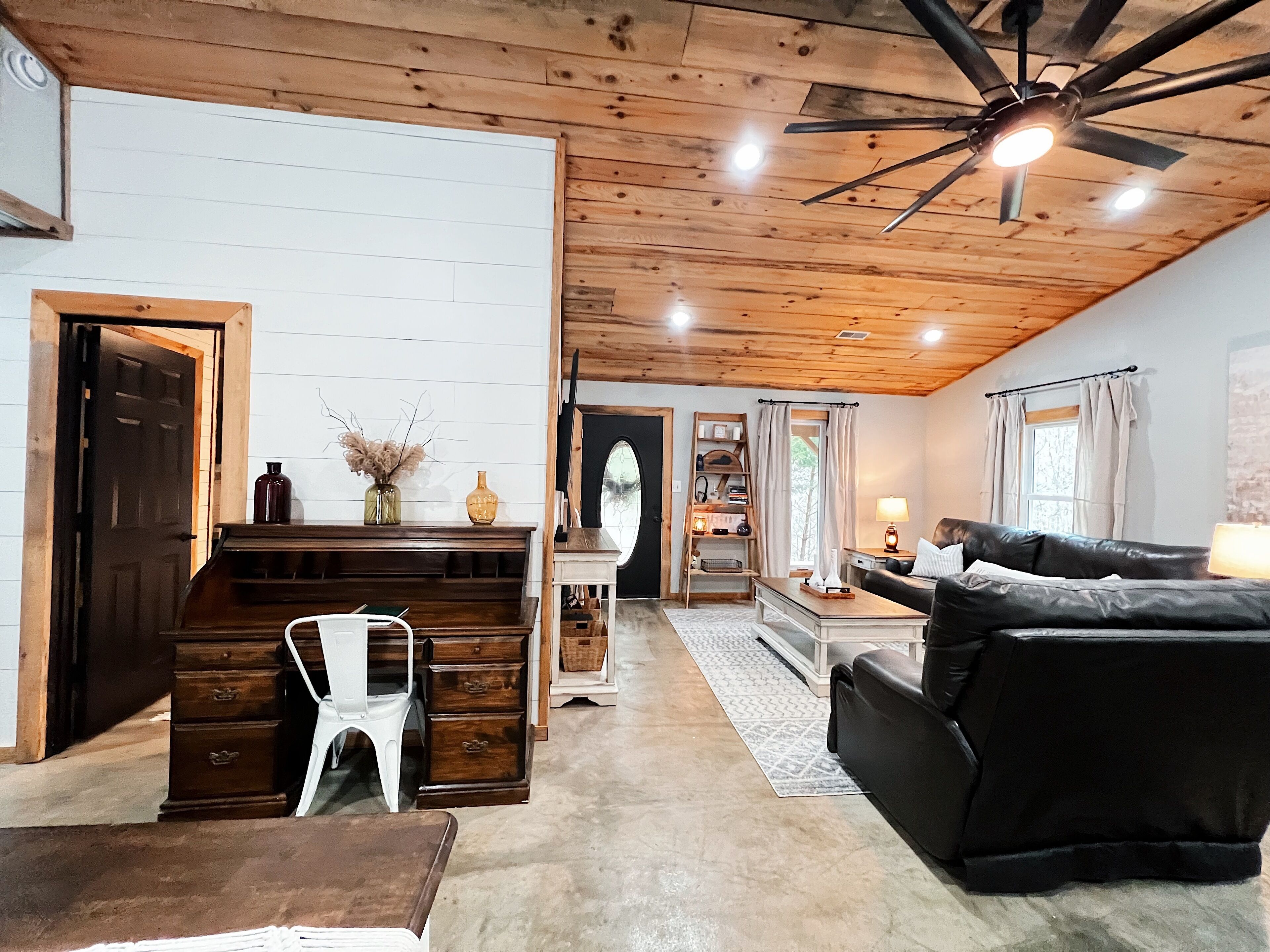 A cozy living room with a wooden ceiling, a black leather sofa, a white chair, and a wooden desk with a vase of dried flowers.