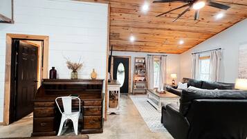 A cozy living room with a wooden ceiling, a black leather sofa, a white chair, and a wooden desk with a vase of dried flowers.