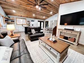 A spacious living room with a leather sofa, a black armchair, and a wooden coffee table. A television is mounted on the wall, and there is a kitchen area in the background.