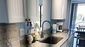 A kitchen with white cabinets, a granite countertop, a sink, and a coffee maker. - Next to the Beach - Our House at the Beach W-103 (Siesta Key)