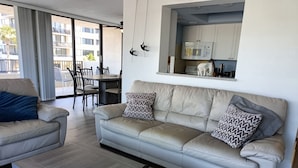 A modern living room with a beige sofa, two patterned pillows, a wooden coffee table, and a kitchen area with a microwave and utensils. - Next to the Beach - Our House at the Beach W-103 (Siesta Key)