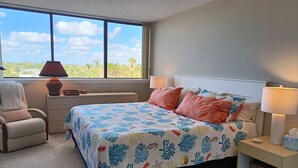 Quarto com uma cama grande, duas luminárias de cabeceira, mesa de cabeceira, uma cadeira e janela com vista para o verde.