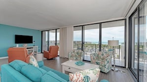 A modern living room with a large window, a flat-screen TV, and a balcony with a view of the ocean.