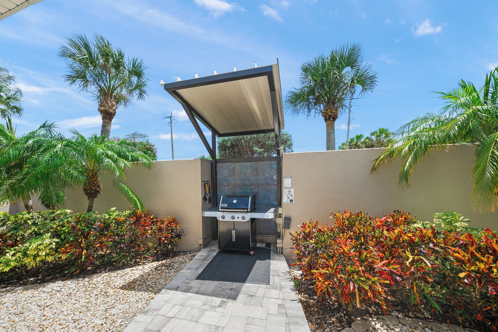 An outdoor grill area with a canopy, surrounded by palm trees and colorful plants.